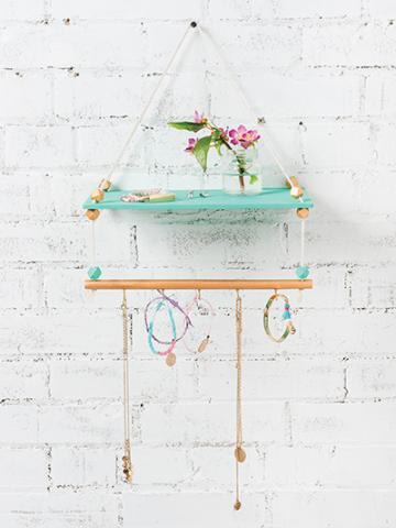a white wall with a hanging shelf in front. the top shelf is teal, the bottom shelf is tan. there are pieces of jewelry hanging from the bottom and a pink flower on the top shelf.