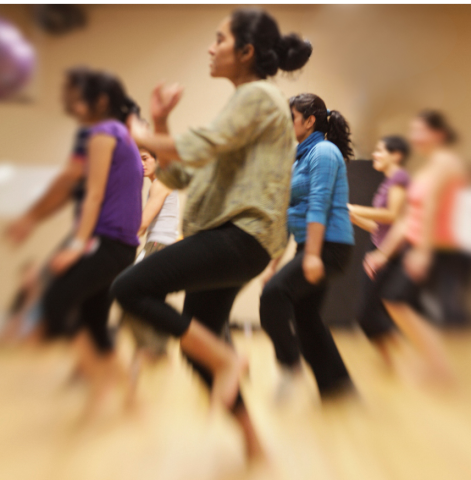 A photo of a a diverse group of dancers in activewear caught in various stages of a dance movement. The dancer clearest and nearest to the front stands with one leg bent and their knee raised as if mid-jog and their arms crossed at the wrists in front of them.