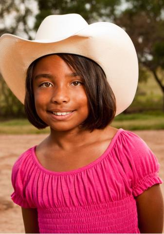 Smiling young brown-skinned girl wearing a pink shirt and a white cowboy hat.