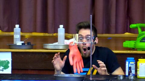 A young white man in a black t-shirt is crouched down behind a table. Surrounding him on small platforms are various types of bottles. He is wearing goggles and his mouth is open in astonishment as he watches the experiment in front of him. A rubbler glove is clipped to a test tube holder in front of him and seems to be unnaturally inflated