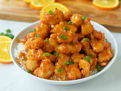 a white bowl with rice and a mound of golden fried orange chicken on a white table with sliced oranges in the background