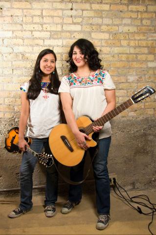 Belinda and Maritza Cervantes, two young Latina women dressed in embroidered shirts and jeans, each holding a guitar.