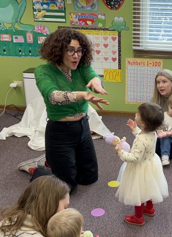 Maestra Claudia, a white woman with curly dark brown hair, kneels on the floor in an early childhood classroom, holding her arm out and interacting with a toddler standing before her while other parents and children can be spotted on the periphery looking on.