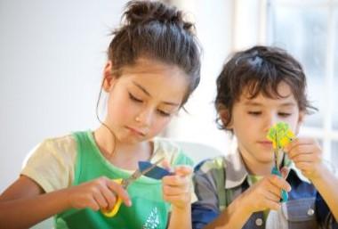 Photo of two dark haired children, a young girl and a slightly younger boy crafting using child scissors.