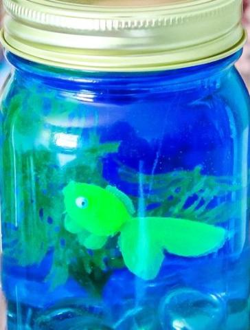 Close up photo of a mason jar containing a little greenery, blue liquid, and a little green toy fish