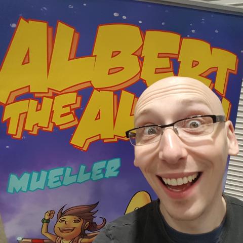 A photo of the artist and author Trevor Mueller, a youthful looking white man with a shaved head and glasses. He is smiling widely and stands in front of an illustration of his creation Albert the Alien