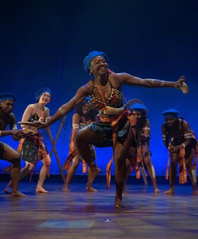 A photo of one of Ayodele Drum and Dance's dance performances. A central dancer dances exuberantly, arms outstretched, while four other dancers stand around her leaning forward and seemingly cheering them on.