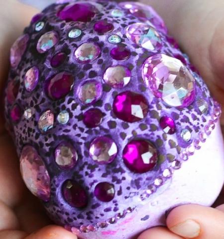 A close-up photo of a person's hand cupped around a large purple egg that is embedded with colorful gems