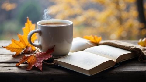 a book is open on a table with a white mug and red leaf against a blurry autumnal background