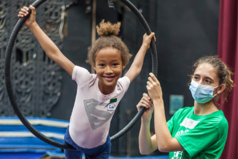 A young light-brown skinned girl smiles as she stretches her arms to grab opposite sides of an oversized ring while a masked CircEsteem trainer steadies the hoop