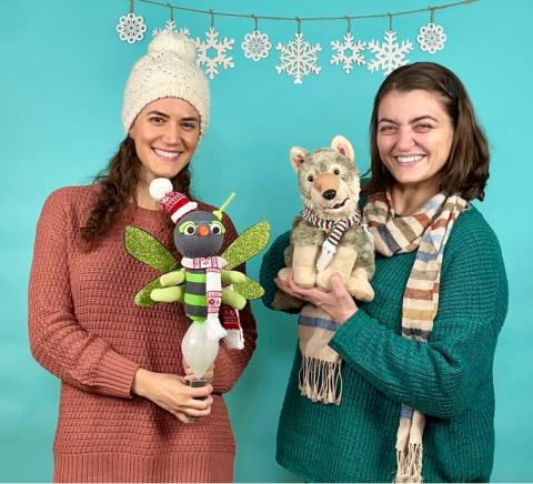 A photo of two of Firefly Family Theatre's performers. They are standing  underneath a delicate snowflake garland and are holding up what appear to be a firefly and wolf puppet