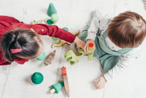 A photo (taken from above) of two young toddlers playing with colored abstract blocks scattered across a pale, wooden floor.