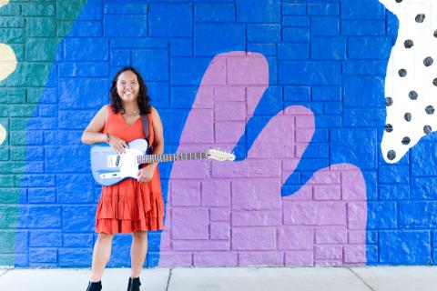 A photo of Filippino-American presenter Little Miss Ann smiling and holding a guitar in front of a wall with an abstract mural.