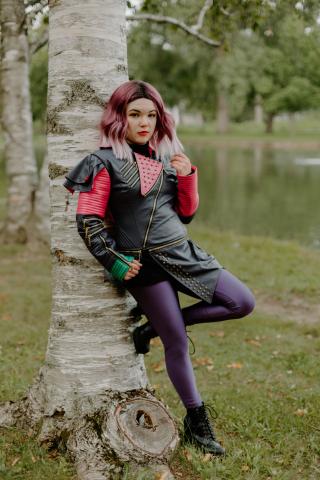 Photo of a young woman with purple ombre hair and tights leaning up against a tree trunk. She is dressed as the Descendant of the Mistress of All Evil.