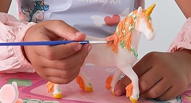 Zoomed in photo image of a childl's hands painting a unicorn figure with a thin paintbrush.