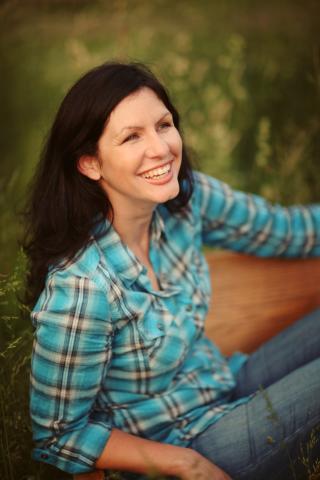 a photo of a white woman with long dark hair wearing a blue plaid shirt, smiling and sitting on a wooden bench with a green background