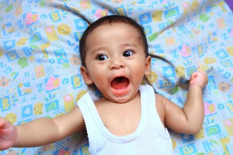 Photo of a light brown-skinned baby with dark hair lying on a baby blanket, arms stretched out and mouth open as if in the middle of a happy exclamation