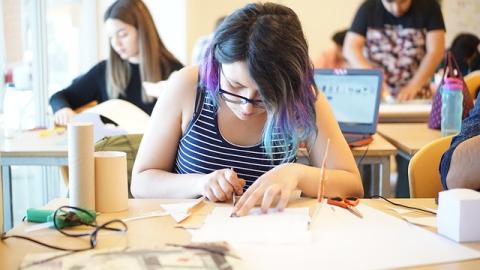 a teen with blue and black hair leaning over a craft project with other teens out of focus in the background