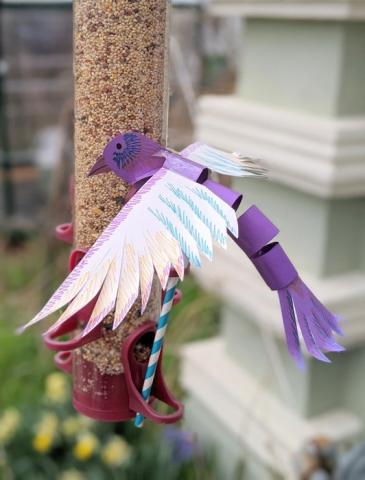 Purple bird made out of paper with decorated white wings. It is attached to a paper straw and next to a bird feeder.