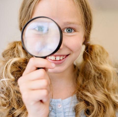 Photo of a young middle grade girl with blonde curly hair in low pigtails. She's holding up a magnifying glass to one eye.
