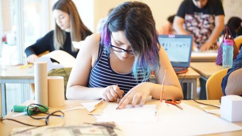 close up shot of a teen with blue hair bent over a crafting table, cutting a piece of paper with other students in the background