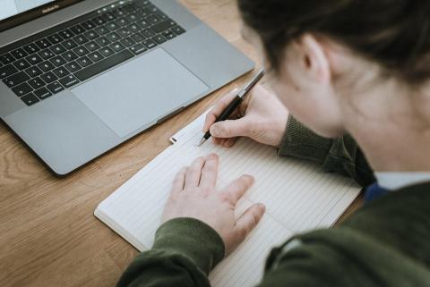 over the shoulder shot of a woman with dark hair writing in a notebook in front of an open laptop