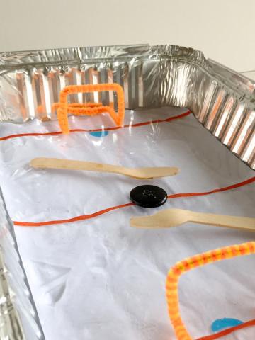 Aluminum pan made to look like an ice hockey rink using pipe cleaners, a button, and popsicle sticks.