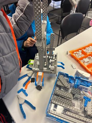 A photo of older kids clustered around a table with a number of Technic-like building pieces before them. They are constructing a tall tower out of the long gray bricks.