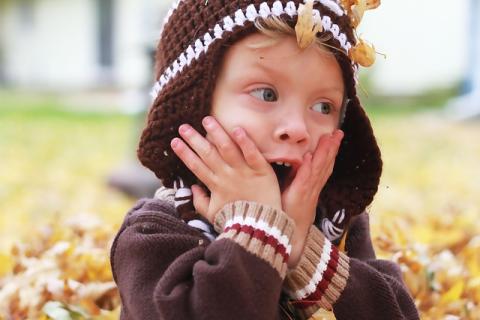 Close-up photo of a white child in a brown fall sweater and matching knit hat. Piles of fall leaves can be seen on the grass behind them, and the child has both of their hands pressed to their face with their mouth open in an expression of happy surprise.