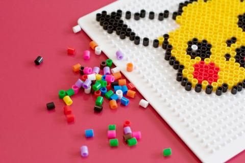 a perler bead version of pikachu on a pink background with loose perler beads next to it