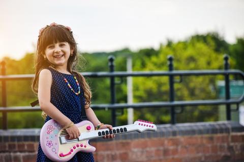 Photo of a little girl with long hair standing outside in front of a fence holding a kid-sized electric guitar