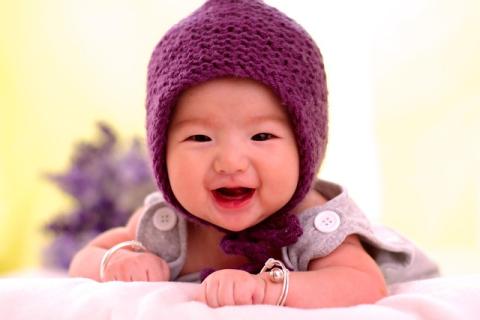 Close up photo of an Asian-presenting baby wearing a knit hat. The baby is lying on their tummy and laughing up at the camera.