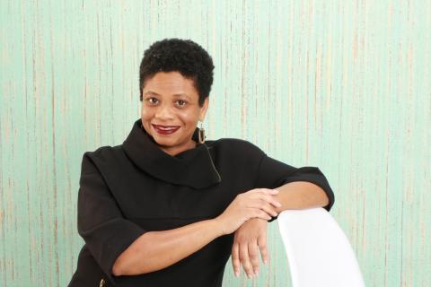 Photo of author Monica Swope, a Black woman with short hair wearing a black sweater. She has her arms gracefully propped on a nearby white chair.