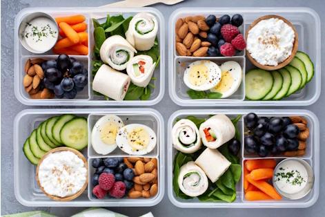 A photo of four variations of bento boxes: small rectangular compartmented containers containing samplings of food such as veggies, nuts, and fruit.