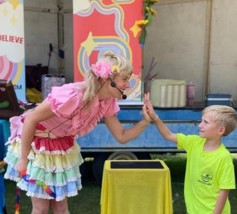 Photo of children's performer Miss Make-Believe, a whimsically-dressed young woman with blonde pigtails, giving a high five to a young boy.s 