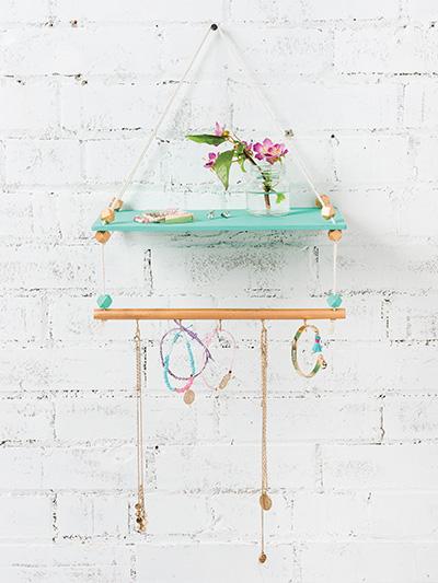 a white wall with a hanging shelf in front. the top shelf is teal, the bottom shelf is tan. there are pieces of jewelry hanging from the bottom and a pink flower on the top shelf.