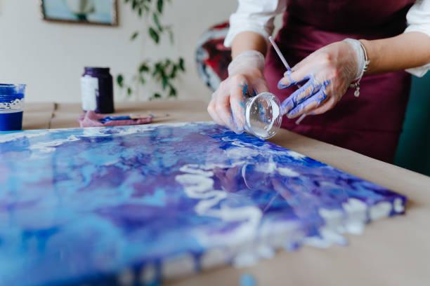 A person wearing gloves and pouring shades of blue and purple paint onto a canvas.