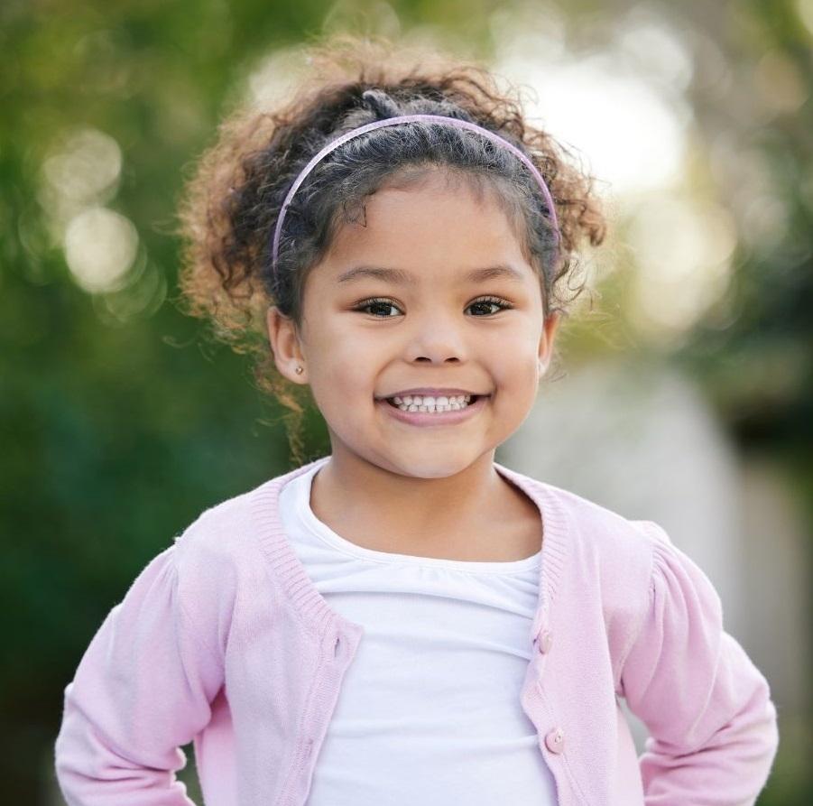 Outside photo with hazy trees in the background. A young black girl stands in the foreground with her hands on her hips. She is smiling widely.