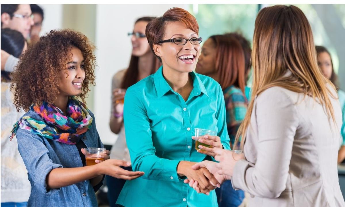 A diverse trio of adult women smile as they greet each other and mingle