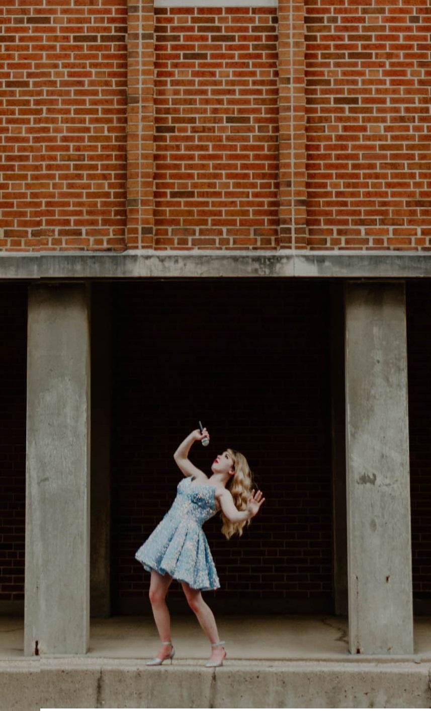 A photo of a young white woman with long wavy blonde hair and wearing a light blue dress, frozen in a position as if she's belting a song, leaning back with a microphone held up in front of her. She stands framed between two pillars of a big brick building.