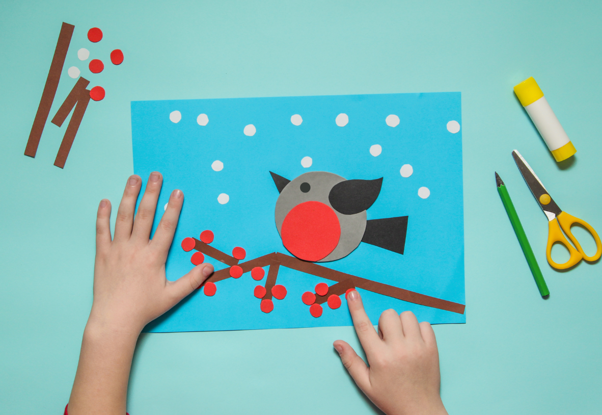 Overhead photo of a child's hands positioning brightly colored construction paper shapes on a blue background to create a snowy bird scene