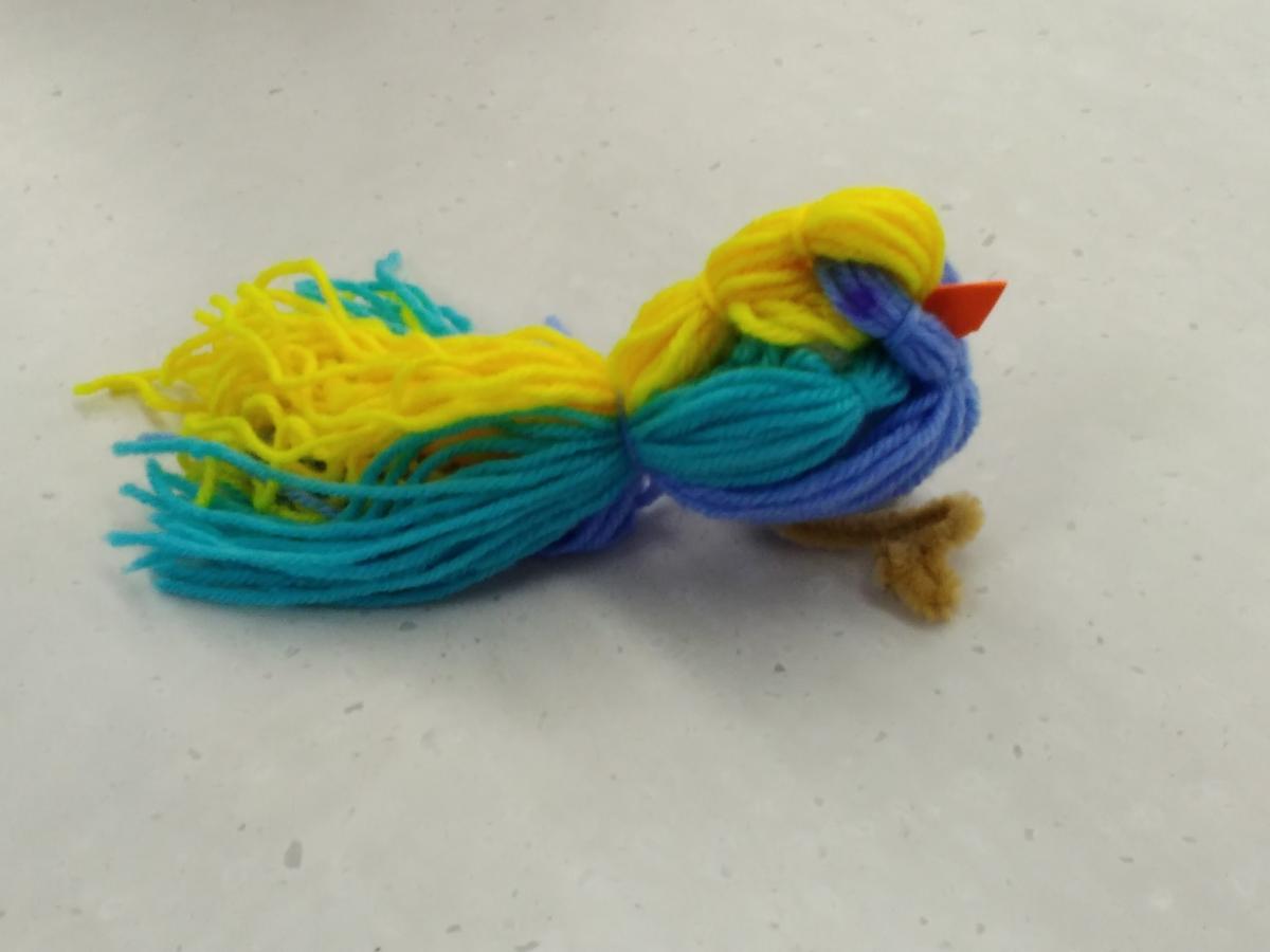 Bird made out of yarn with an orange craft foam beak. The bird is yellow, turquoise, and blue.